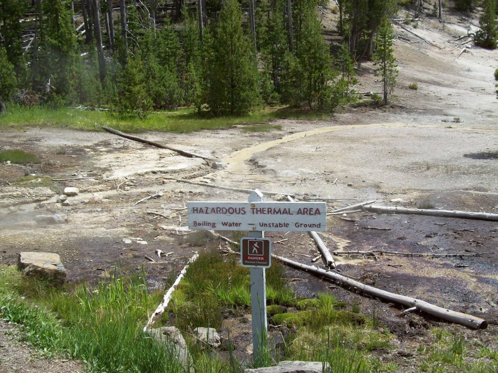 Sign reading Hazardous Geothermal Area. Boiling water. Unstable area. In front of hot spring with trees behind it.