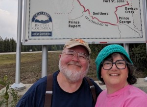 Man and woman standing in front of a sign that says British Columbia. The map shows part of Yellowhead 16 Highway, Prince Rupert and St George.